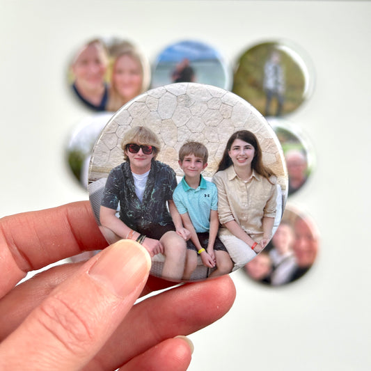 2.25" round button photo magnet being held between two fingers. The magnet shows three children sitting and there are other round photo magnets in the background.